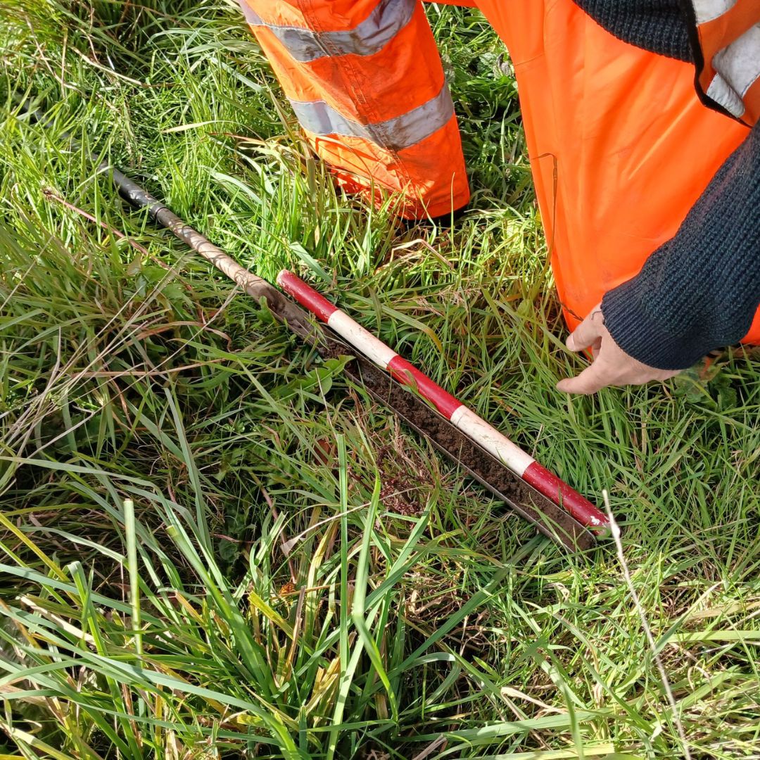 Person wearing bright orange high-visibility work trousers kneeling on grassy ground next to a red and white survey pole and a soil auger. The tools are placed among tall green grass, indicating an outdoor fieldwork or land surveying activity.