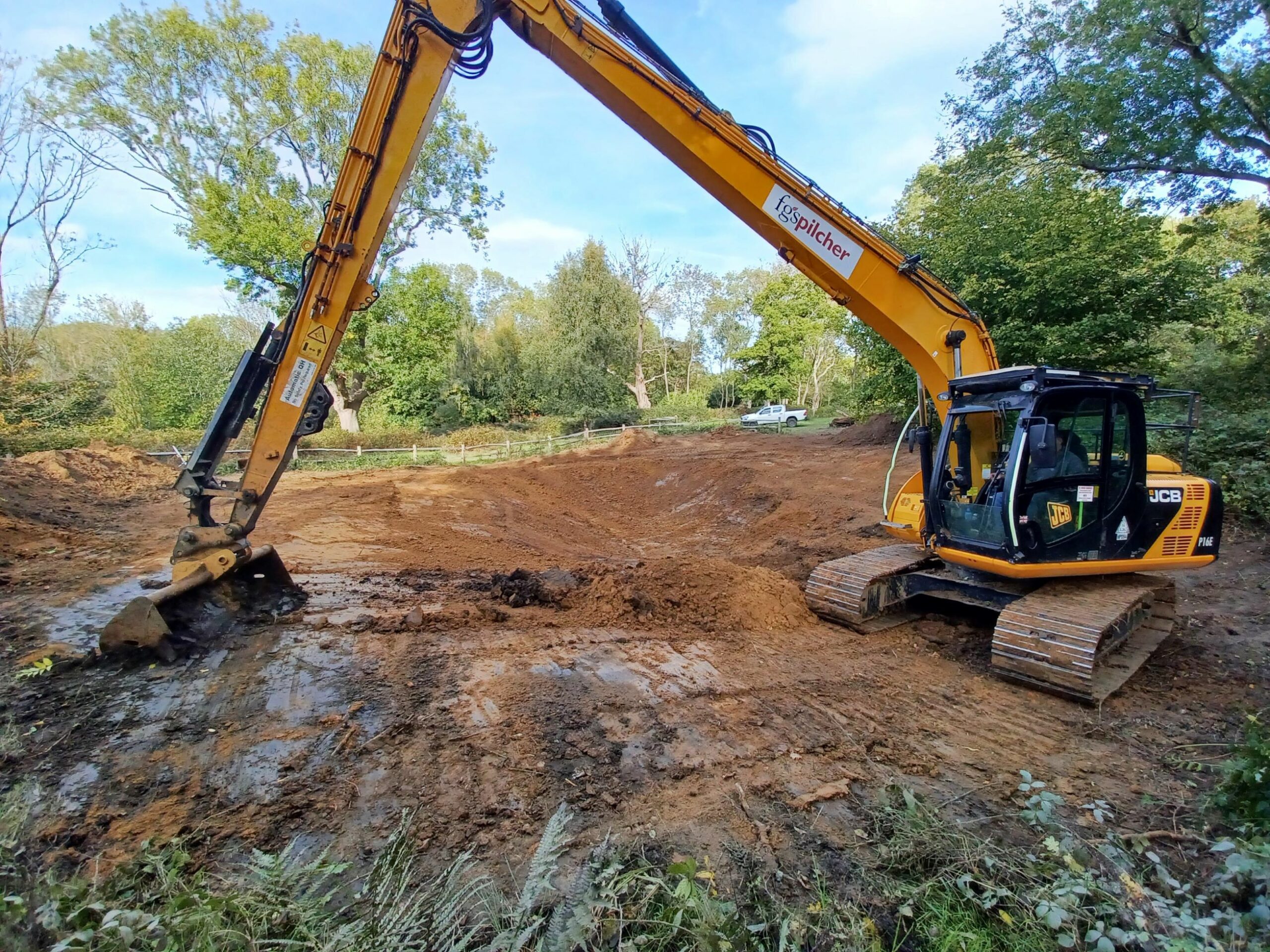Work in progress. Large yellow JCB excavator with extended arm digging and leveling soil on a construction site surrounded by green trees under a clear blue sky.