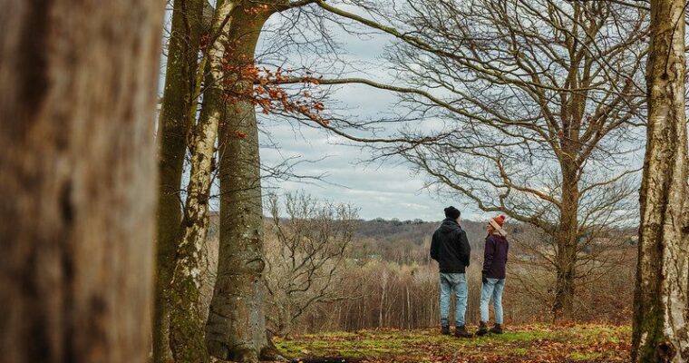 Two people in winter clothes stand chatting in a woodland with views across the valley behind