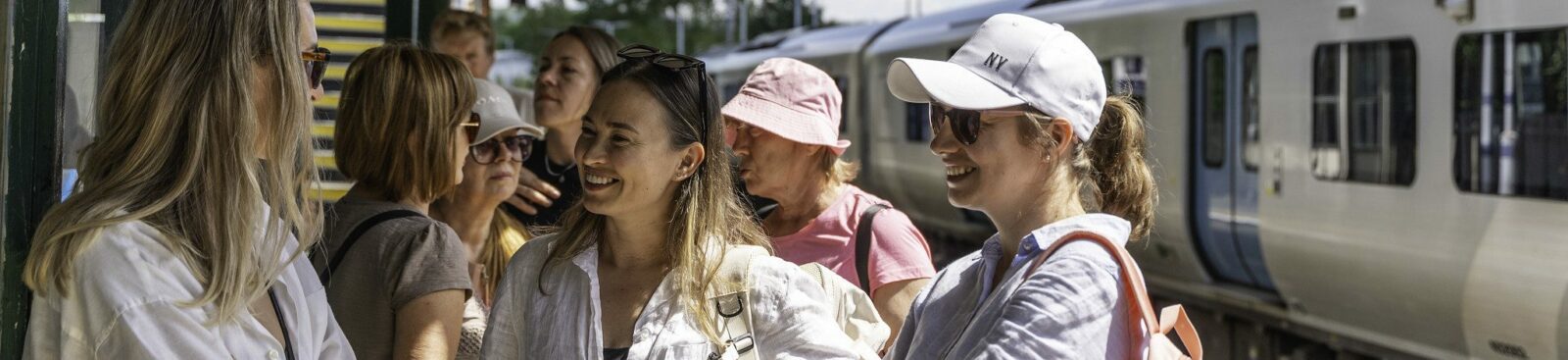 A group of women in summer clothes and sunhats stand on a train platform, with rucksacks to go on a walk