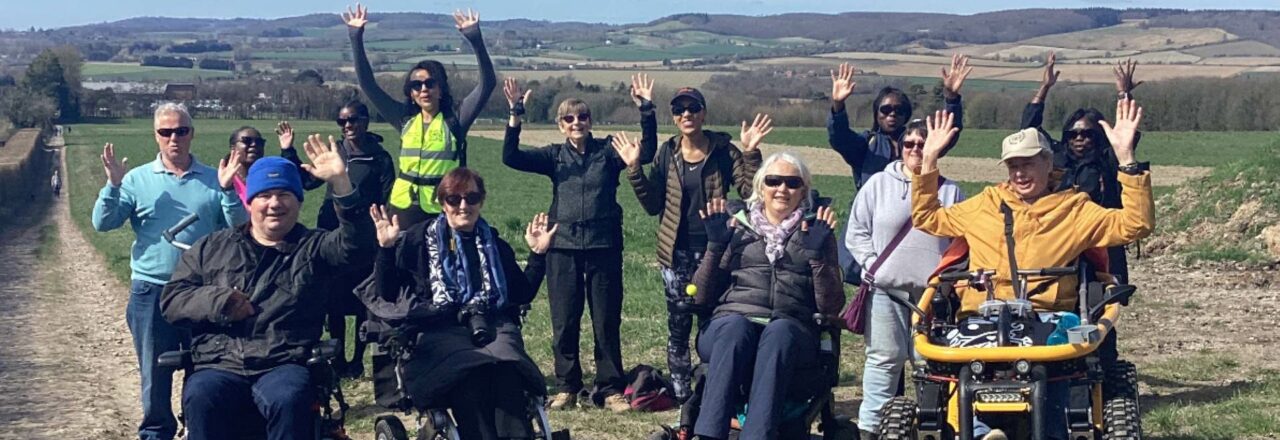 A group of people in wheelchairs raising their hands while on a trail outdoors.