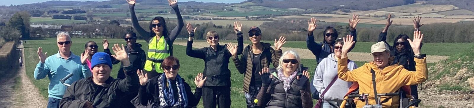A group of people in wheelchairs raising their hands while on a trail outdoors.
