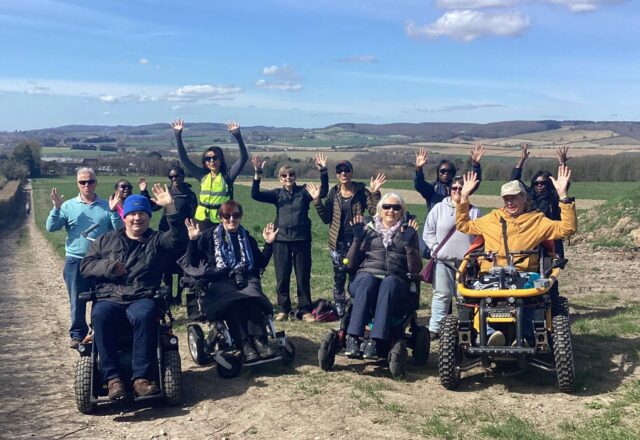 A group of people in wheelchairs raising their hands while on a trail outdoors.