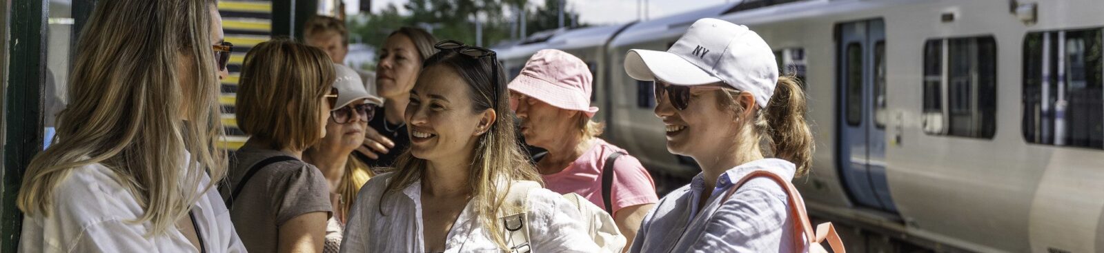 A group of women in summer clothes and sunhats stand on a train platform, with rucksacks to go on a walk