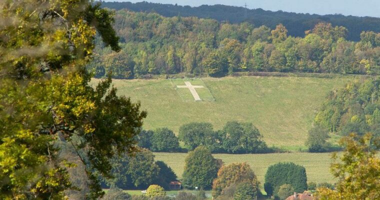 A large white cross stands prominently on a hillside, contrasting with the surrounding green grass. In the foreground, trees with green and yellow leaves frame the scene, suggesting an autumn setting. Below the hill, fields and a small cluster of buildings form a village or town. The background features a dense forest with a mix of green and autumnal hues. The cross, a striking focal point, may hold cultural, historical, or religious significance.